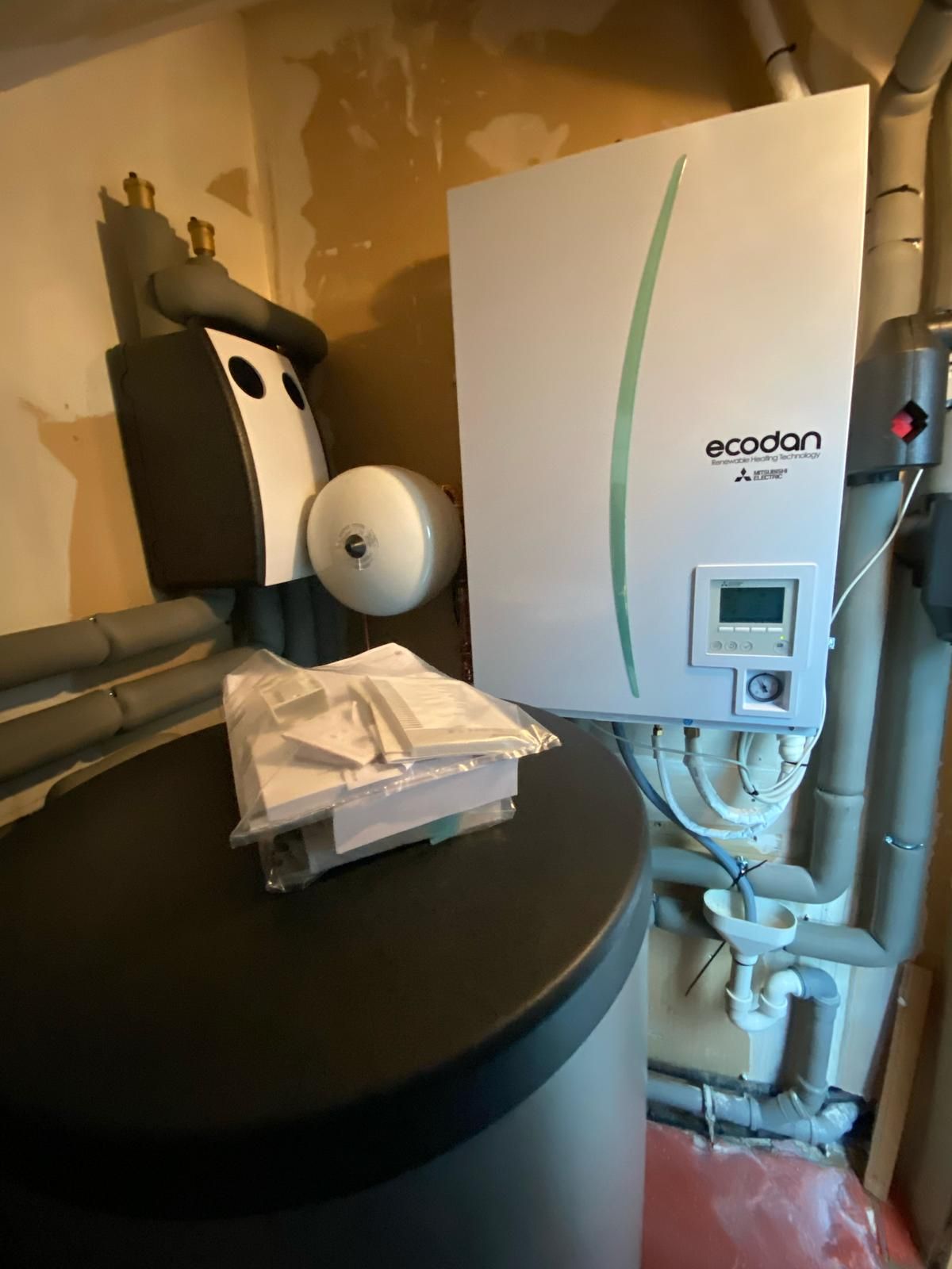Ecodan-Wärmepumpenmodul an Wand mit Pufferspeicher, Isolierungen und aufliegenden Bedienungsanleitungen.