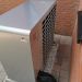 Rückseite einer Wärmepumpe mit Lüftungsgitter und Isolierschlauch, auf Kiesbett zwischen zwei Ziegelwänden.