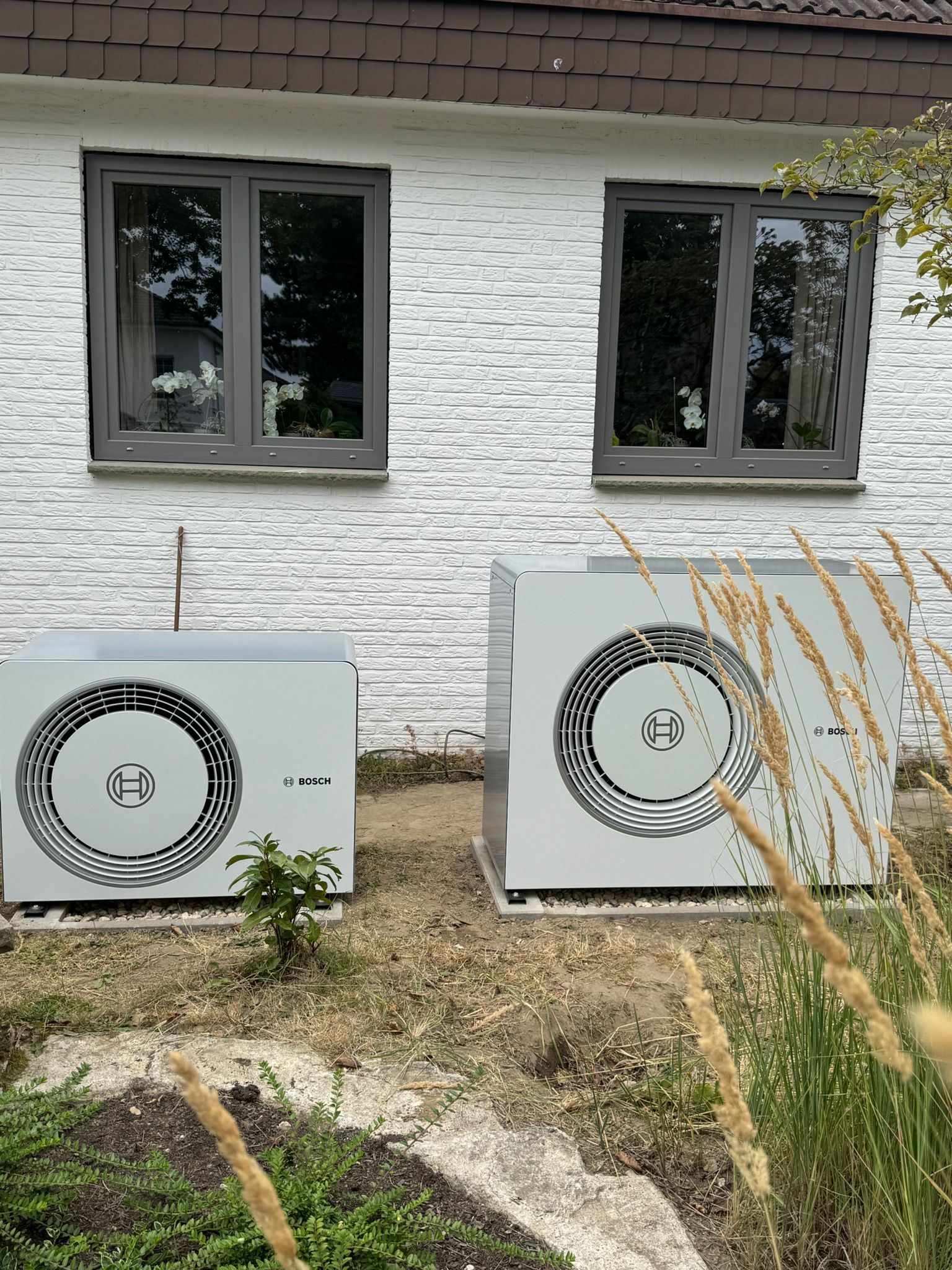 Zwei Bosch Wärmepumpen Außeneinheiten vor weißer Hausfassade im Gartenbereich.