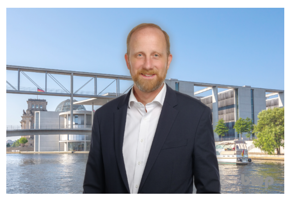 Ein Mann im Anzug steht vor dem Berliner Regierungsviertel mit Spree, Reichstag und moderner Brücke im Hintergrund.