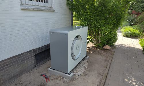 Bosch-Wärmepumpe außen auf Kiesbett vor weißer Hauswand, neben Gehweg und Sträuchern, mit Werkzeug im Vordergrund.