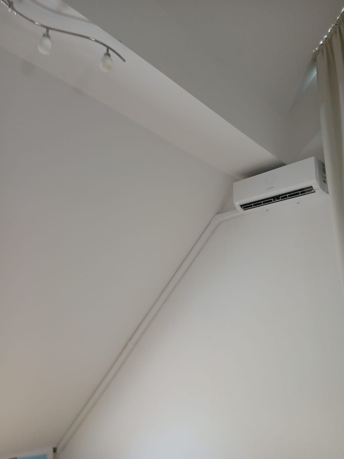 Innenklimagerät an schräger Decke mit sichtbarer Verrohrung und Deckenleuchte im weißen Zimmer.