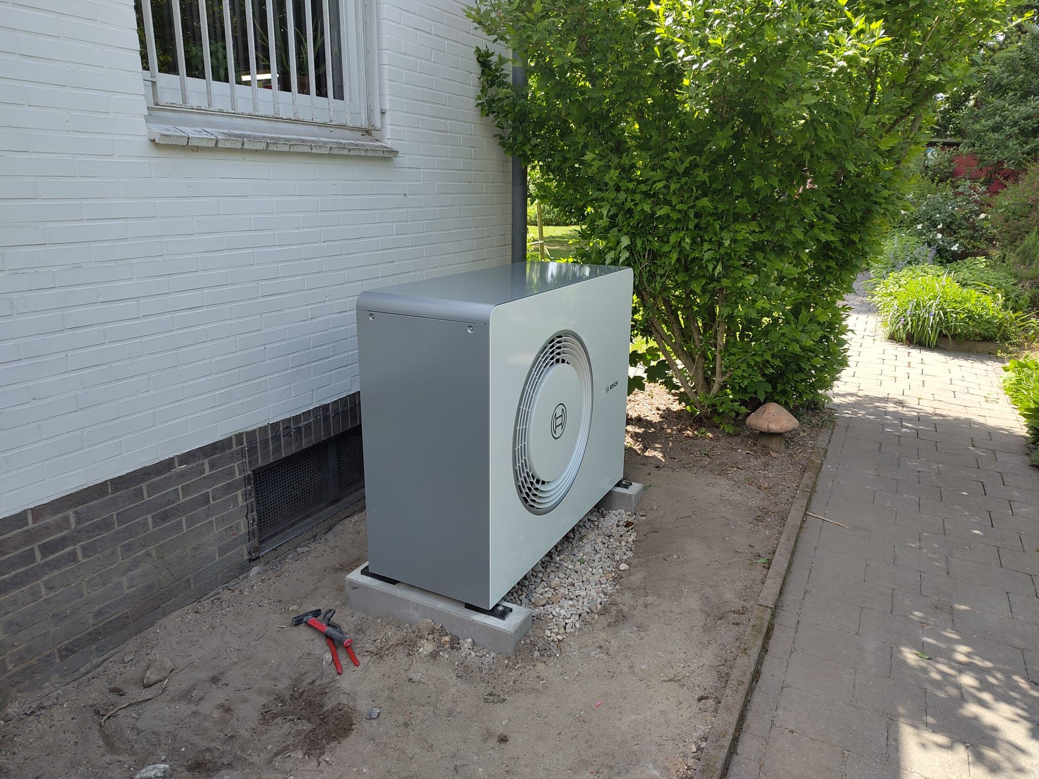 Bosch-Wärmepumpe außen auf Kiesbett vor weißer Hauswand, neben Gehweg und Sträuchern, mit Werkzeug im Vordergrund.