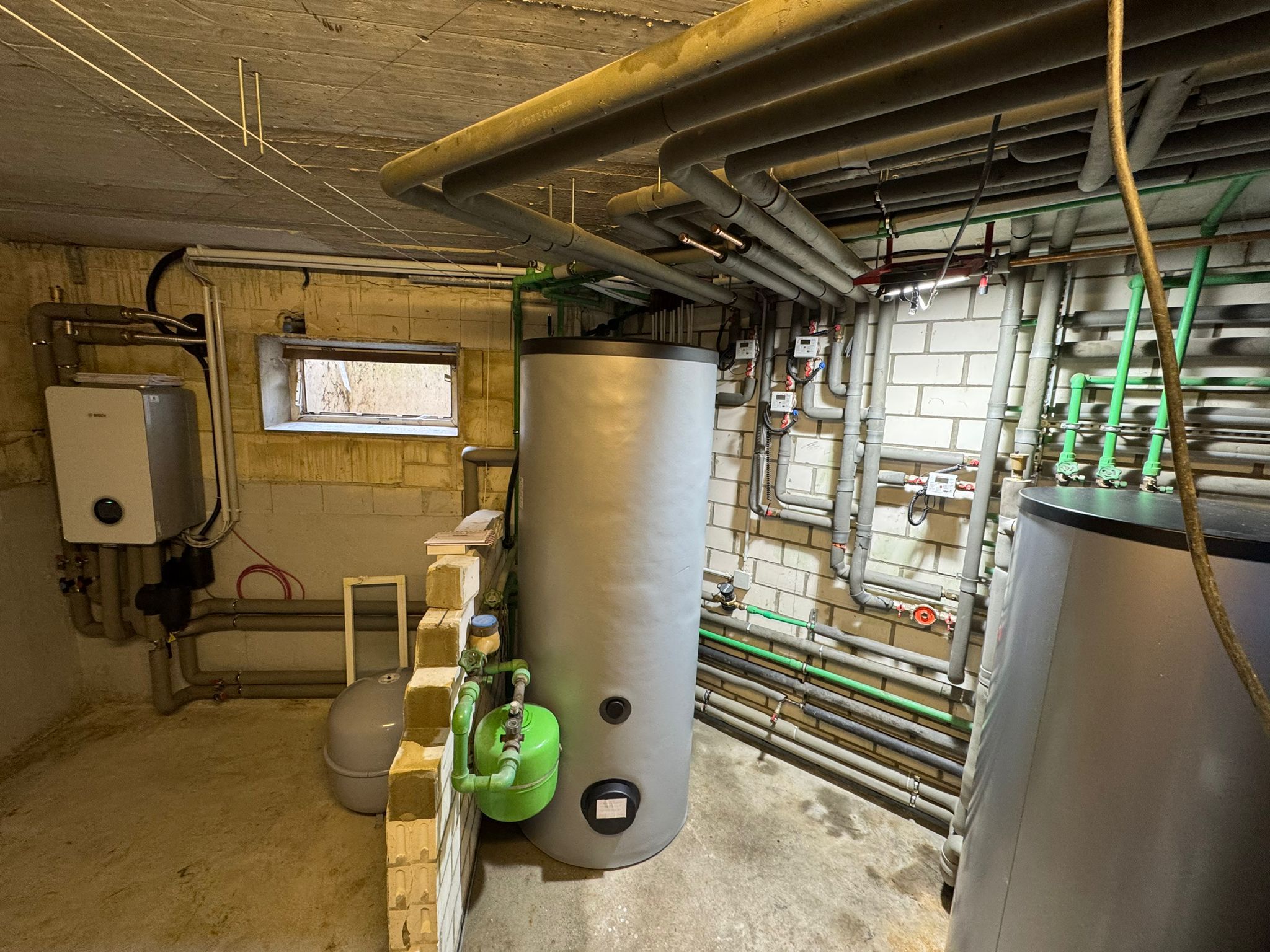Kellerraum mit großem Pufferspeicher, Rohrleitungen und Bosch Inneneinheit an Wand.