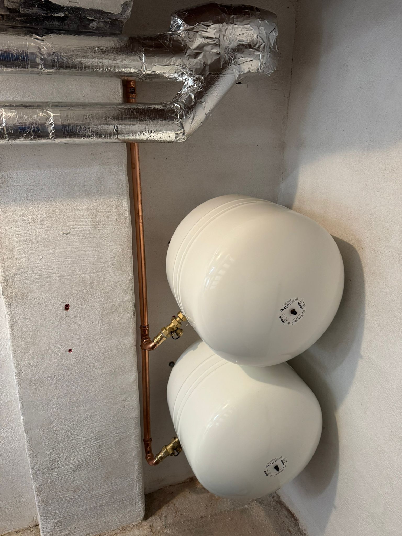 Zwei weiße Ausdehnungsgefäße an Kellerwand, angeschlossen an Heizungsrohre.